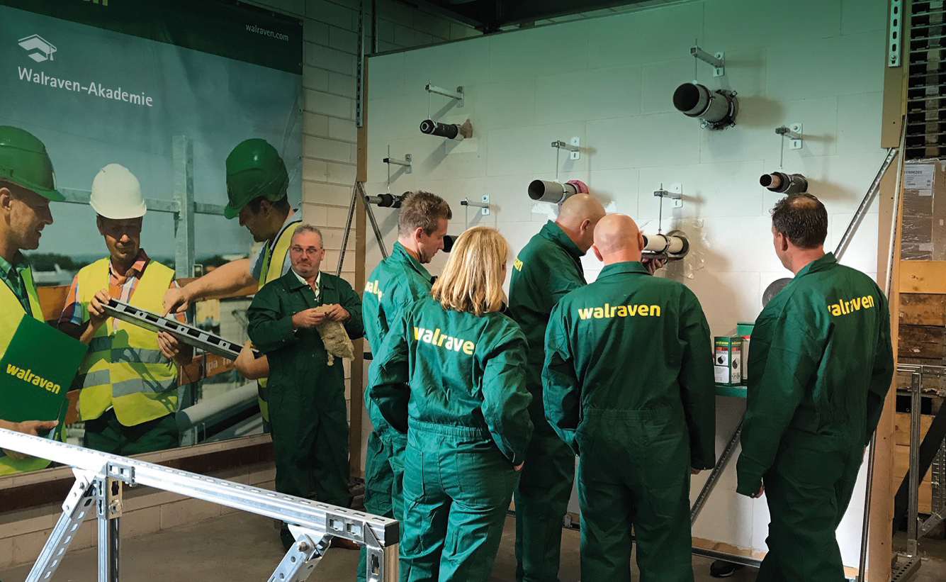 Fünf Menschen in Walraven-Overalls vor einer Wand mit Musterrohrabschottungen. Links davon ein Plakat mit drei Monteuren mit Helmen und einer Montageschiene, darüber ein stilisierter Doktorhut mit dem Schriftzug "Walraven-Akademie".