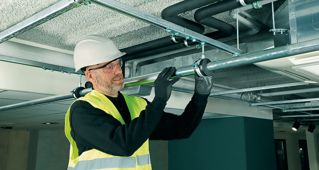 Ein Mann befestigt eine Walraven Kälterohrschelle an einem Rohr. Im Hintergrund ist eine Zwischendecke zu sehen.