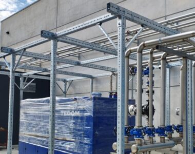 Die installierte Schwerlast-Lösung von Walraven für RTS Srl: Eine maßgeschneiderte Rahmenkonstruktion aus Metallprofilen trägt sicher eine große, blau verpackte Maschine sowie eine komplexe Verrohrung aus Edelstahl mit blauen Ventilen.