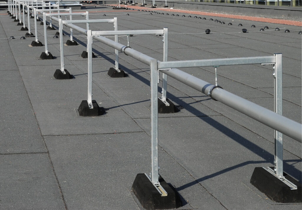 Auf einem Flachdach verlaufen mehrere Rohrleitungen, die auf metallischen Montageschienensystemen mit schwarzen Dämpfungssockeln montiert sind. Die Sockel sorgen für eine stabile, schwingungsdämpfende Aufständerung ohne Dachdurchdringung.