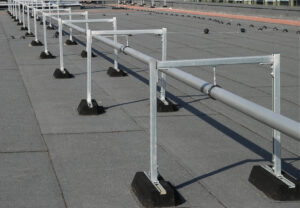 Auf einem Flachdach verlaufen mehrere Rohrleitungen, die auf metallischen Montageschienensystemen mit schwarzen Dämpfungssockeln montiert sind. Die Sockel sorgen für eine stabile, schwingungsdämpfende Aufständerung ohne Dachdurchdringung.