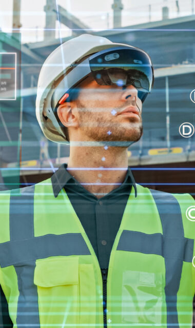 Futuristic Architectural Engineer Wearing Augmented Reality Headset, Uses Gestures to Create 3D Graphics VFX Model of a Building with Infographics. In Background Construction Site in Progress